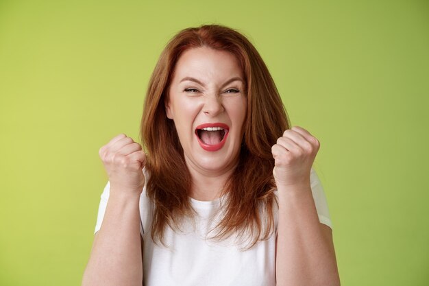 Sim, conseguimos, feliz, feliz, ruiva, de meia-idade, vencedora do sexo feminino, levanta os punhos celebração gesto de sucesso gritando triunfo alegria sorrindo amplamente comemorar incrível banca de jornal parede verde feliz