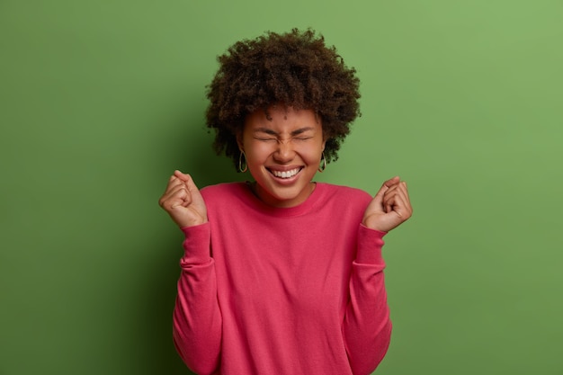 Sim, esta é minha oportunidade. mulher afro-americana alegre regozija-se com o sucesso, fecha os punhos em triunfo, deseja vencer, celebra a conquista, veste um suéter rosado, isolada na parede verde