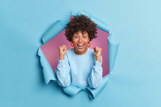 Sim, finalmente consegui. Jovem excitada de pele escura com cabelo encaracolado cerrando os punhos celebrando algo incrível com um sorriso amplo