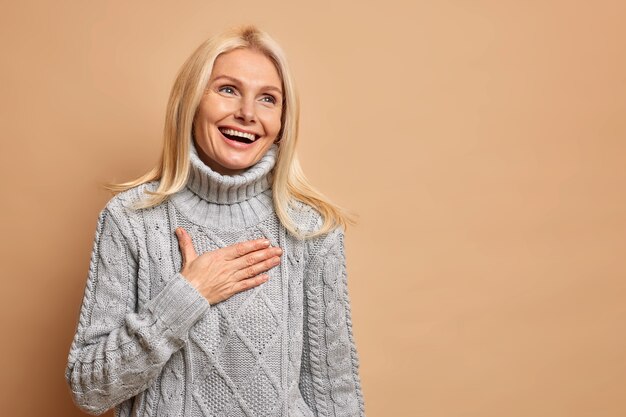 Sincera positiva mulher de meia-idade ri feliz mantém a mão no peito sorri amplamente tem pele saudável, maquiagem mínima reage a algo agradável usa suéter cinza.