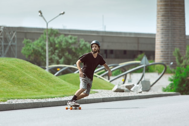 Skatista fazendo uma manobra na rua da cidade em dia ensolarado. Jovem em equipamento de equitação e longboard no asfalto em ação. Conceito de atividade de lazer, esporte, radical, hobby e movimento.