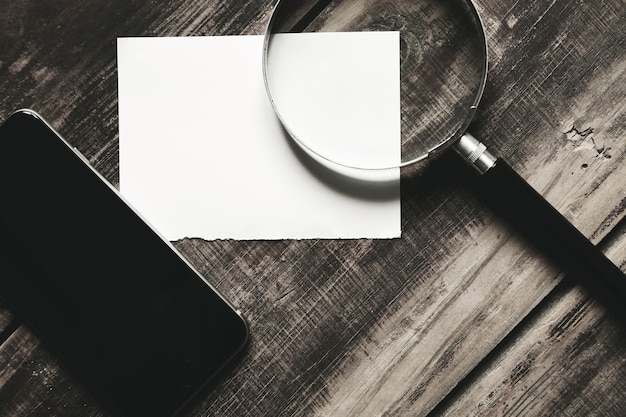 Smartphone móvel, lupa e folha de papel branco isoladas na mesa de madeira de fazenda negra Conceito de jogo de detetive misterioso. Vista lateral do close up