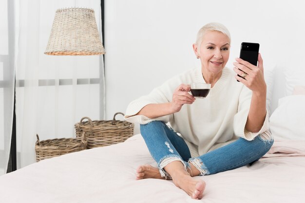 Smiley mulher idosa segurando a xícara de café e olhando para o telefone na cama