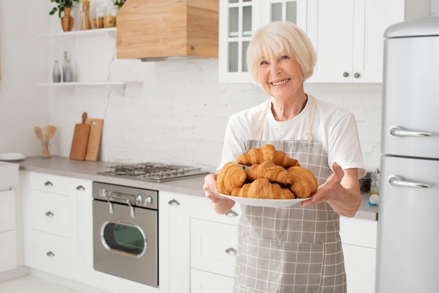 Foto grátis smiley velha segurando um prato com croissants na cozinha