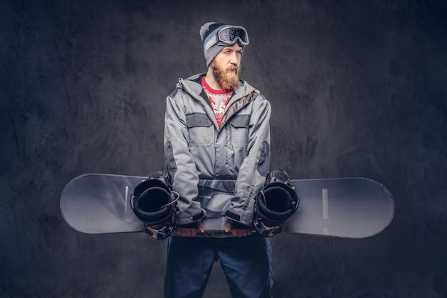 Foto grátis snowboarder barbudo ruivo com um chapéu de inverno e óculos de proteção vestido com um casaco de snowboard segura uma prancha de snowboard em um estúdio. isolado em um plano de fundo texturizado escuro.