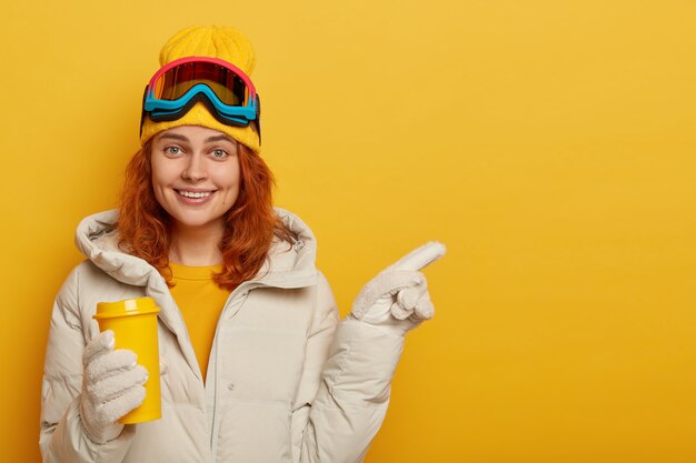 Snowboarder mulher adulta com cabelo ruivo, gosta de bebida quente no inverno, usa roupas de esqui, aponta para um espaço livre para seu conteúdo promocional ou texto.