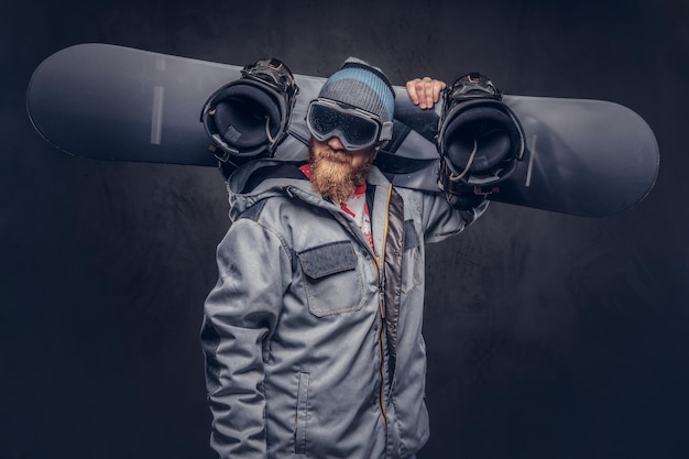 Snowboarder ruiva brutal com uma barba cheia em um chapéu de inverno e óculos de proteção vestido com um casaco de snowboard segura uma prancha de snowboard no ombro em um estúdio. Isolado em fundo cinza.