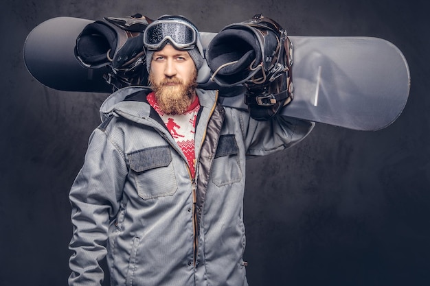 Snowboarder ruiva brutal com uma barba cheia em um chapéu de inverno e óculos de proteção vestido com um casaco de snowboard segura uma prancha de snowboard no ombro em um estúdio. Isolado em fundo cinza.