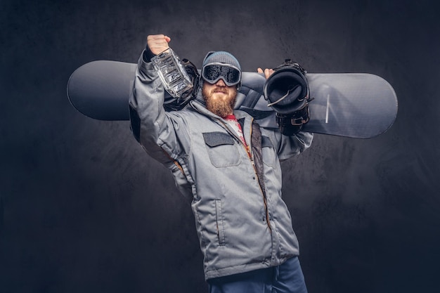 Snowboarder ruiva com uma barba cheia em um chapéu de inverno e óculos de proteção vestido com um casaco de snowboard segura uma prancha de snowboard e uma garrafa de álcool no ombro em um estúdio. Isolado nas costas cinza