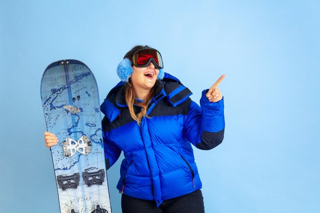Snowboarding. Retrato de uma mulher caucasiana sobre fundo azul do estúdio. Linda modelo feminino com roupas quentes. Conceito de emoções, expressão facial, vendas, anúncio. Clima de inverno, época de Natal, feriados.