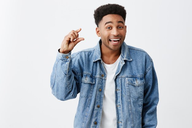 Só um pouco. Feche o retrato isolado de jovem bonito de pele negra com penteado afro em camiseta branca sob jaqueta jeans azul gesticulando com a mão, olhando na câmera com feliz