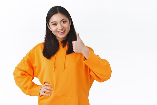 Soa bem. Mulher asiática otimista otimista com retrato da cintura para cima usando um capuz laranja, mostrando o polegar para cima em gesto de aprovação e sorrindo com a cabeça em aceitação