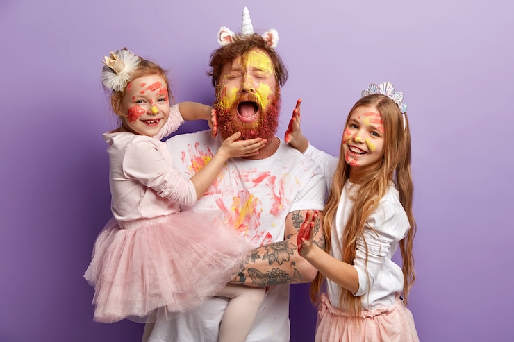 Foto grátis sobrecarregado o cansaço, pai solteiro com barba ruiva, chora desesperadamente, se diverte com duas meninas, usa tintas coloridas, tem expressões alegres, fica de pé sobre a parede roxa. conceito de feliz dia dos pais