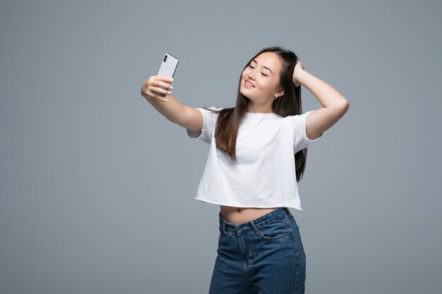 Sociável menina asiática linda tomando selfie ou falando na vídeo chamada usando telefone celular sobre fundo cinza