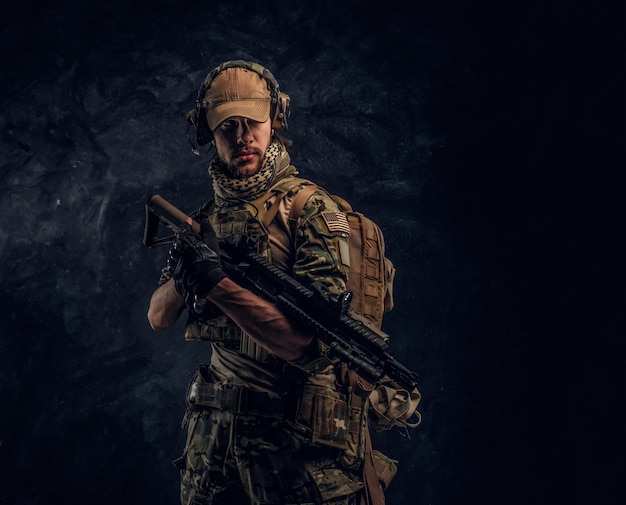 Soldado totalmente equipado em uniforme de camuflagem segurando um rifle de assalto. Foto de estúdio contra uma parede texturizada escura