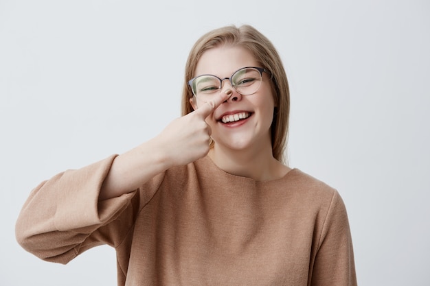 Sombria jovem caucasiana zomba, toca o nariz, zomba, ri e sorri amplamente, demonstra seus dentes brancos e uniformes. A menina loura positiva sorri alegremente