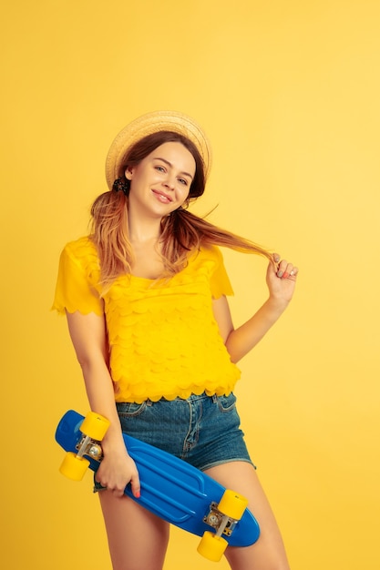 Sonhadora, segura o skate. Retrato de uma mulher caucasiana em fundo amarelo do estúdio. Bela modelo feminino no chapéu. Conceito de emoções humanas, expressão facial, vendas, anúncio. Verão, viagens, resort.