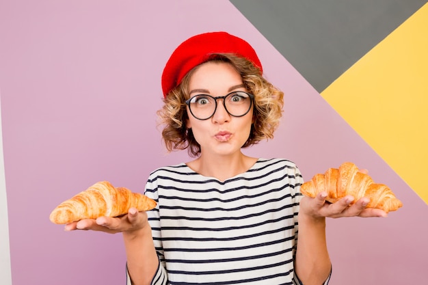 Sonhadora senhora bonita na boina vermelha segurando grandes croissants saborosos nas mãos.