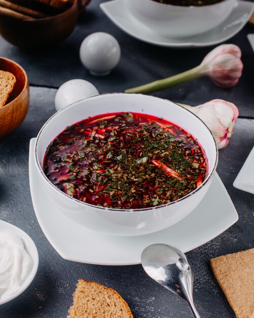 sopa quente de borsh com legumes cozidos dentro de um prato branco redondo com ovos de pão na mesa cinza