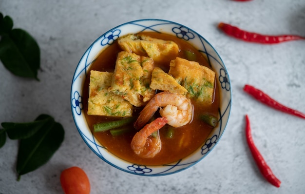 Sopa quente e azeda com cha-om, ovo e camarão em uma tigela branca, com pimenta e folhas de lima kaffir na superfície branca.