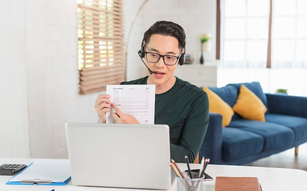 Sorridente bonito empresário asiático usa fones de ouvido trabalhando remotamente de casa. Ele é uma videoconferência webinar