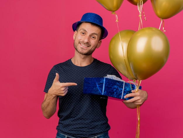 Foto grátis sorridente, bonito homem caucasiano com chapéu de festa azul aponta e aponta para balões de hélio isolados em um fundo rosa com espaço de cópia