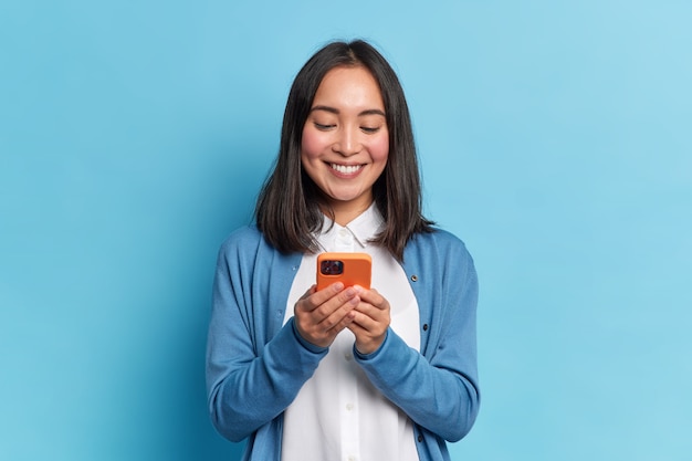 Sorridente e charmosa mulher asiática morena usa celular feliz mensagens de texto em redes sociais viciada em tecnologias modernas usa blusão casual