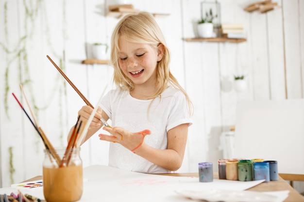 Sorridente e feliz menina loira de camiseta branca, desenhando algo na palma da mão com um pincel