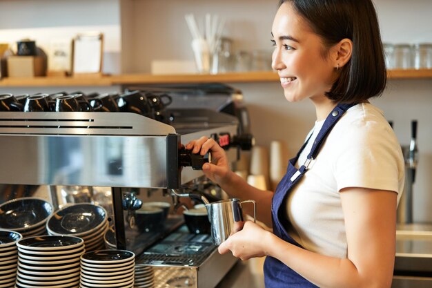 Sorridente jovem barista asiática usando máquina de café vaporizando leite para pedido de cappuccino em pé no avental trabalhando no café