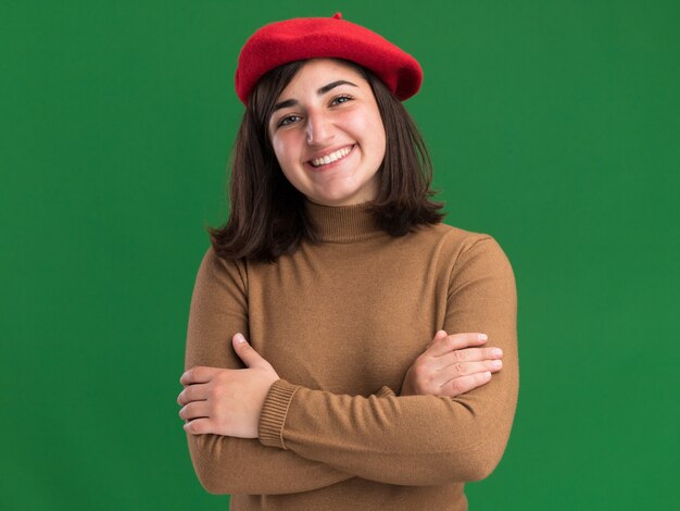 Sorridente, jovem, bonita, caucasiana, com chapéu boina fica com os braços cruzados no verde