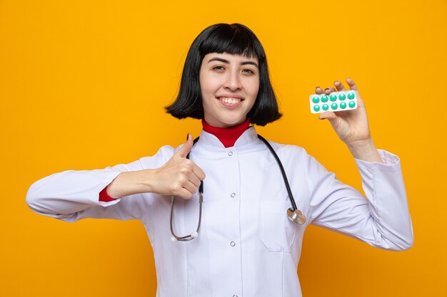 Sorridente, jovem, bonita, caucasiana, com, uniforme médico, com, estetoscópio, segurando, embalagem, pílula, e, thumbing up