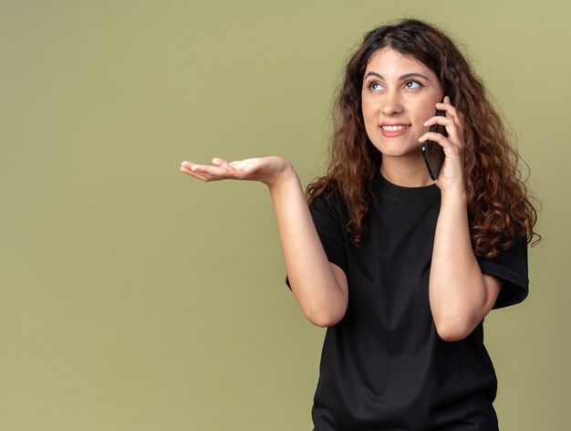 sorridente jovem bonita caucasiana falando no telefone olhando para cima, mostrando a mão vazia isolada na parede verde oliva com espaço de cópia