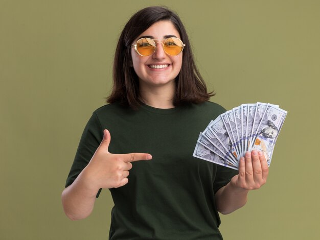 Sorridente, jovem, bonita, caucasiana, usando óculos de sol, segurando e apontando para o dinheiro, olhando para a câmera no verde oliva