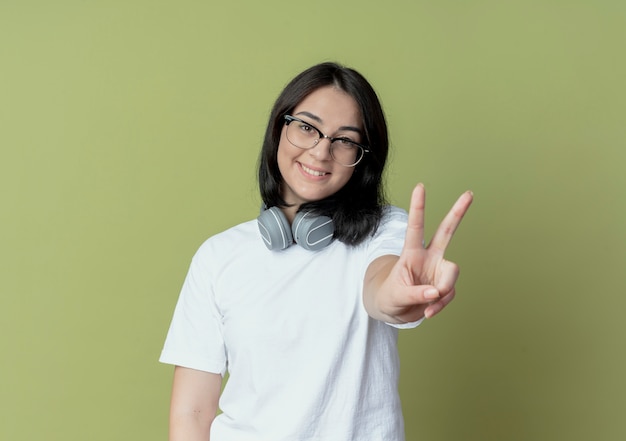 Sorridente, jovem, bonita, caucasiana, usando óculos e fones de ouvido no pescoço, fazendo o sinal da paz, isolado em um fundo verde oliva com espaço de cópia