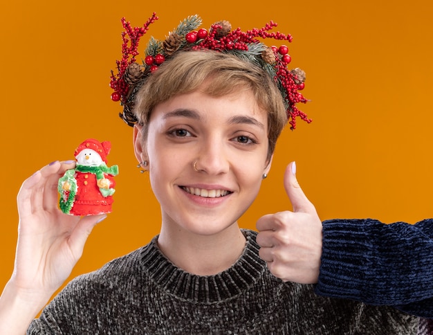 sorridente jovem bonita usando coroa de natal segurando uma pequena estátua de boneco de neve de natal mostrando o polegar isolado na parede laranja