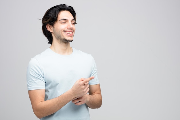 sorridente jovem bonito apontando para o lado com os olhos fechados, isolados no fundo branco com espaço de cópia