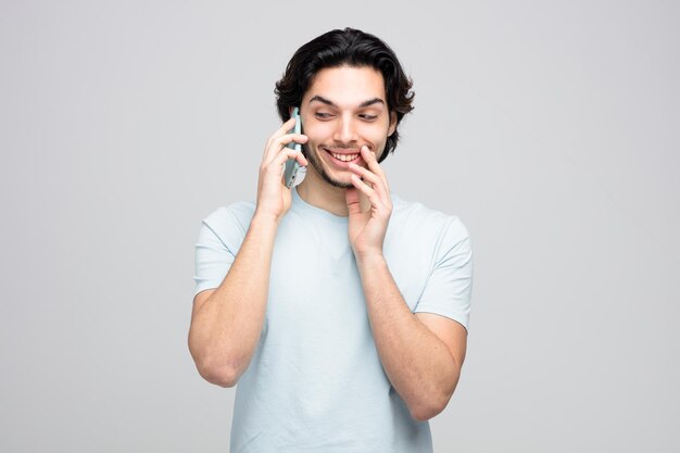 sorridente jovem bonito falando no telefone, mantendo a mão perto da boca, olhando para o lado sussurrando isolado no fundo branco