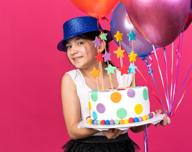 sorridente jovem caucasiana com chapéu de festa azul segurando um bolo de aniversário e balões de hélio isolados na parede rosa com espaço de cópia