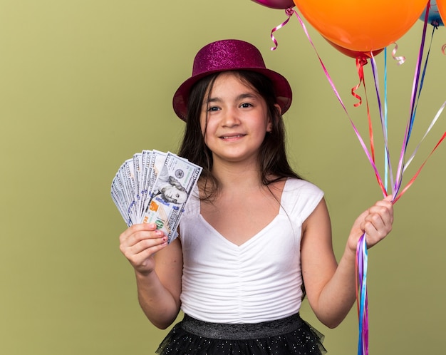 sorridente jovem caucasiana com chapéu de festa roxo segurando dinheiro e balões de hélio isolados na parede verde oliva com espaço de cópia