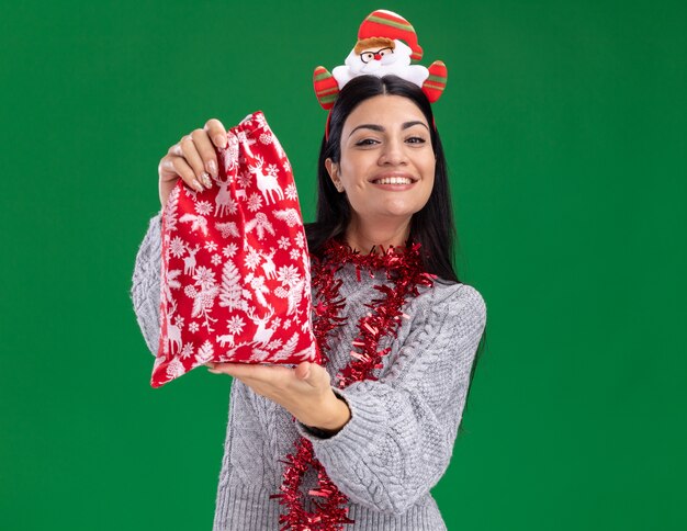 sorridente, jovem, caucasiana, vestindo uma faixa de papai noel e guirlanda de ouropel no pescoço, estendendo o saco de presente de Natal para isolado na parede verde com espaço de cópia