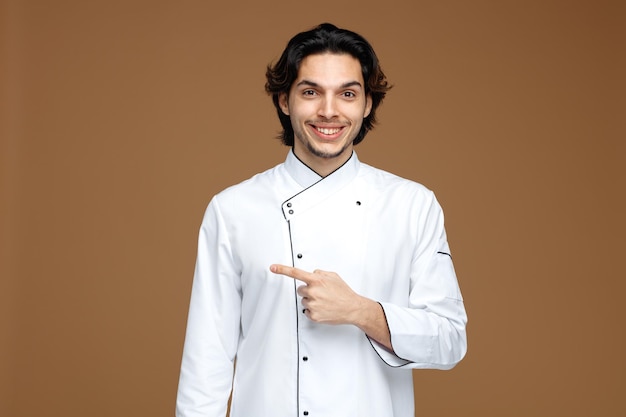 sorridente jovem chef masculino vestindo uniforme olhando para a câmera apontando para o lado isolado no fundo marrom