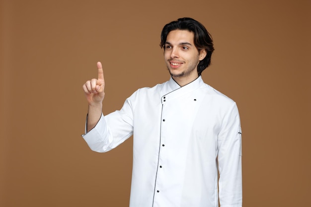 Sorridente jovem chef masculino vestindo uniforme olhando para o lado apontando para cima isolado no fundo marrom