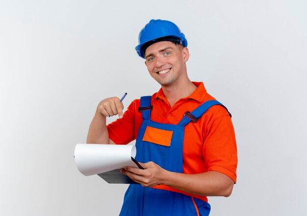 sorridente jovem construtor masculino vestindo uniforme e segurando capacete de segurança e aponta para a área de transferência