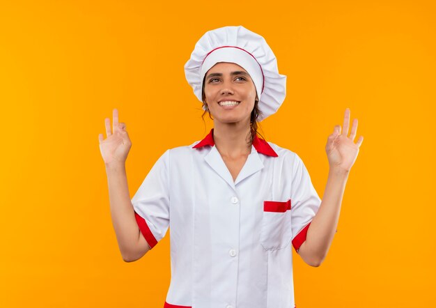 sorridente jovem cozinheira vestindo uniforme de chef, mostrando gesto ok na parede amarela isolada com espaço de cópia