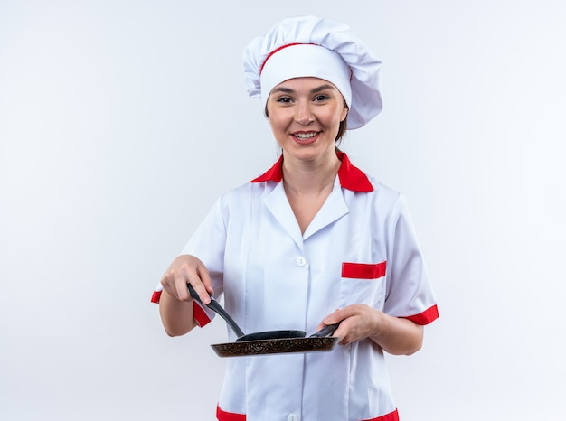 sorridente jovem cozinheira vestindo uniforme de chef segurando uma frigideira com espátula isolada na parede branca