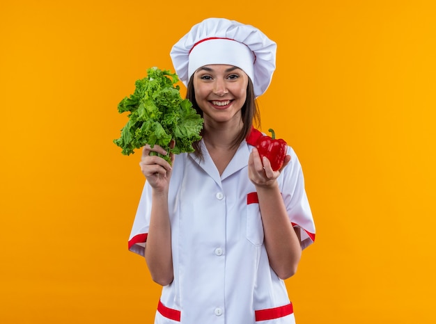 sorridente jovem cozinheira vestindo uniforme de chef segurando uma salada com pimenta isolada na parede laranja