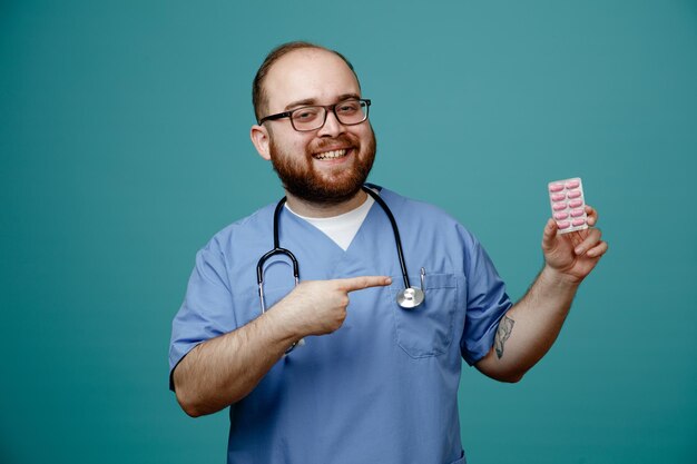 sorridente jovem enfermeiro usando óculos esfoliante de enfermeira e estetoscópio em volta do pescoço, olhando para a câmera mostrando o pacote de cápsulas apontando para ele isolado no fundo azul