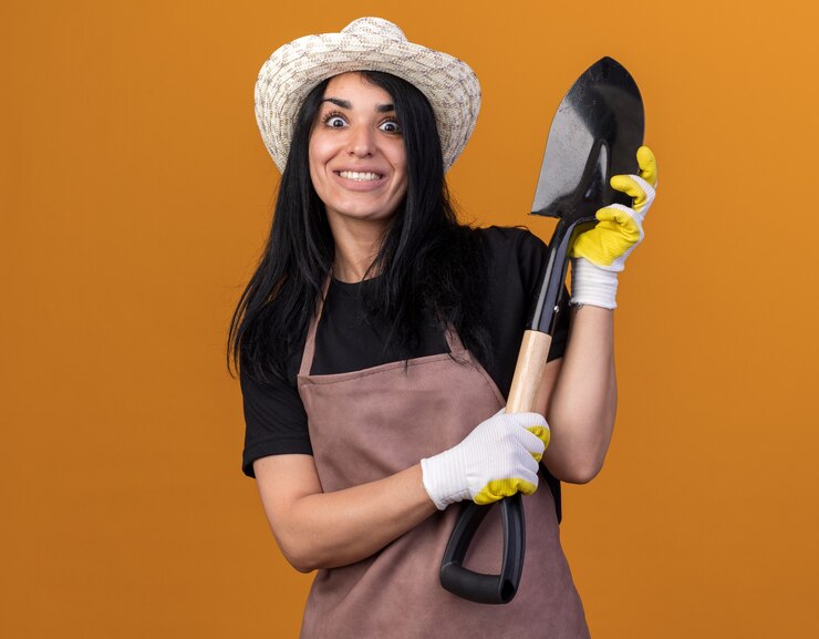 Foto grátis sorridente jovem jardineira caucasiana vestindo uniforme e chapéu com luvas de jardineiro segurando uma pá isolada na parede laranja com espaço de cópia