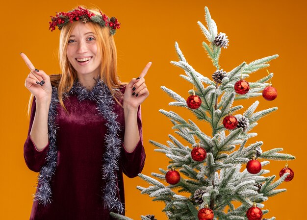 sorridente jovem linda em pé perto da árvore de Natal com vestido vermelho e grinalda com guirlanda no pescoço aponta para cima isolado na parede laranja