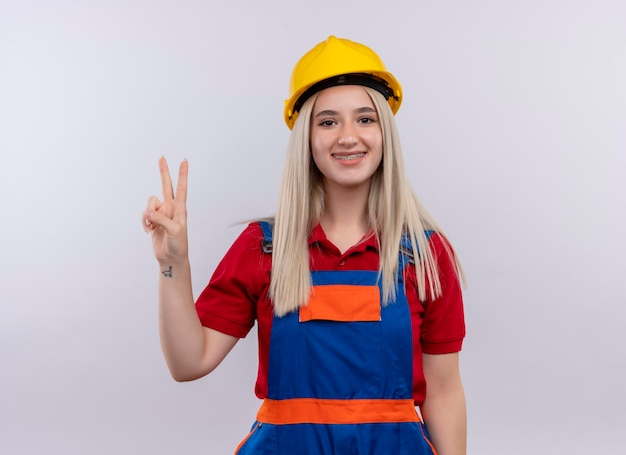 Sorridente jovem loira engenheira construtora de uniforme com aparelho dentário fazendo sinal da paz em um espaço em branco isolado